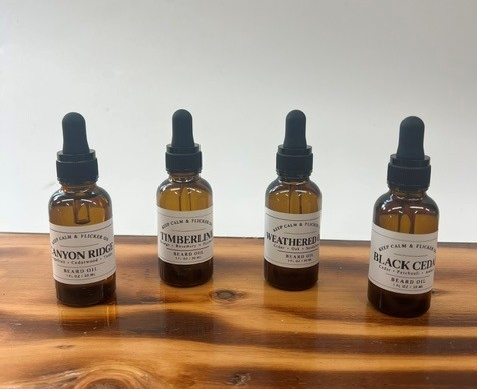 Four amber glass dropper bottles of beard oil arranged on a wooden surface against a plain white background. The labels read Canyon Ridge, Timberline, Weathered, and Black Cedar.