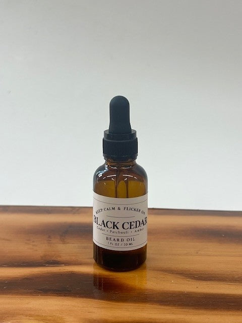 A small amber glass dropper bottle of Black Cedar Beard Oil with a black dropper cap and white label, standing on a wooden surface against a plain light background.