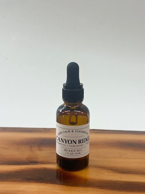 A 1 fl oz amber glass dropper bottle of Canyon Ridge Beard Oil with a black rubber dropper cap, resting on a wooden surface against a light gray background.